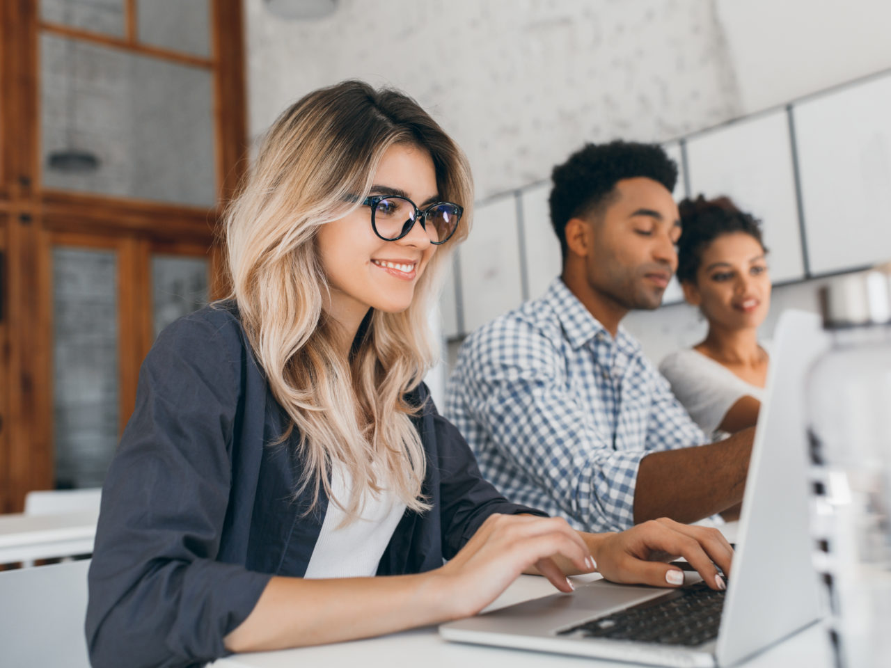 Beautiful curly female freelancer with cute manicure using laptop and smiling. Indoor portrait of blonde secretary sitting beside african coworker in blue shirt.